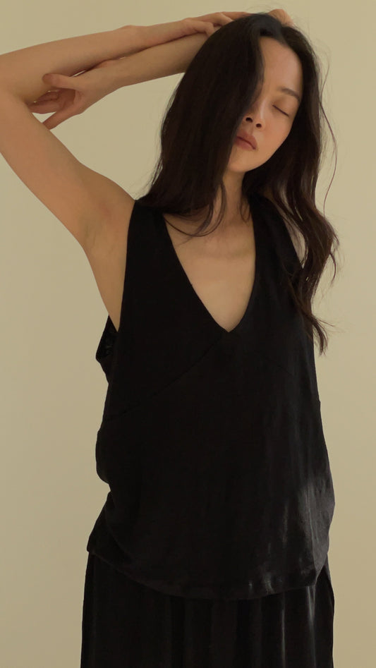 A person with long dark hair, eyes closed, poses with one arm raised above their head in a Soft Tank against a plain light background.