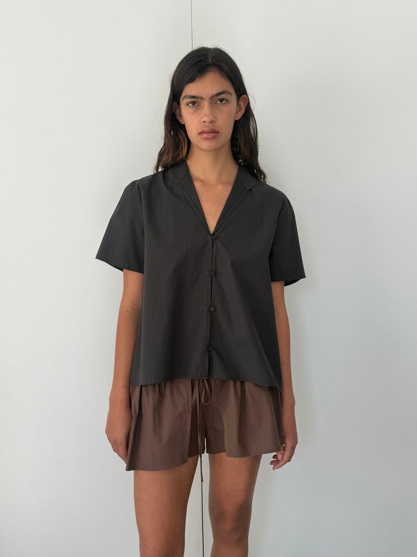 A woman with long dark hair stands against a plain white wall, wearing the Sails Top in Black Stripe and loose brown shorts. She looks directly at the camera with a neutral expression.