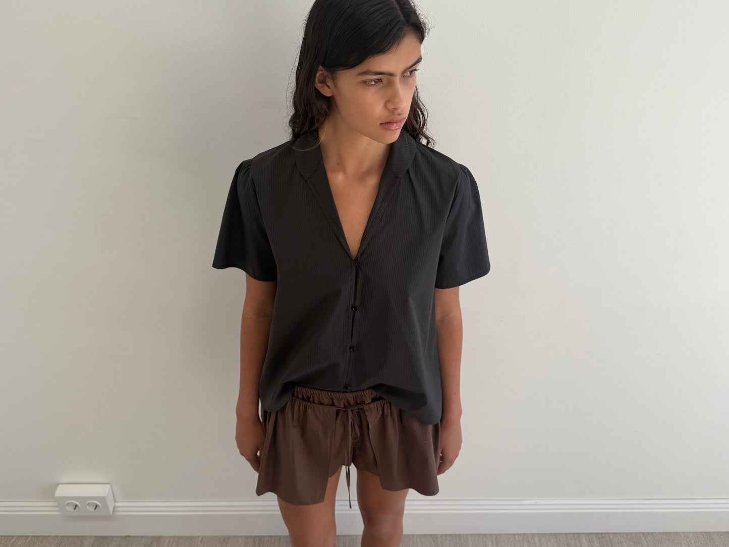 A person with long dark hair stands indoors against a plain white wall, wearing the Sails Top - Black Stripe, an oversized short-sleeved shirt made from organic cotton, paired with brown shorts, looking slightly to the right.