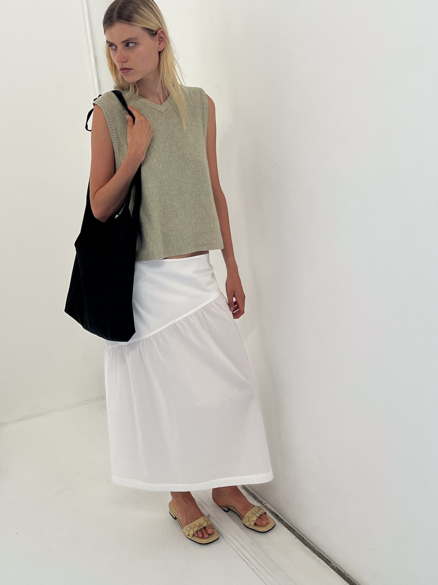 A woman with straight light blonde hair and a neutral expression stands against a white wall wearing the Angle Skirt - White, a sleeveless beige top, sandals, and carrying a large black tote bag over one shoulder.