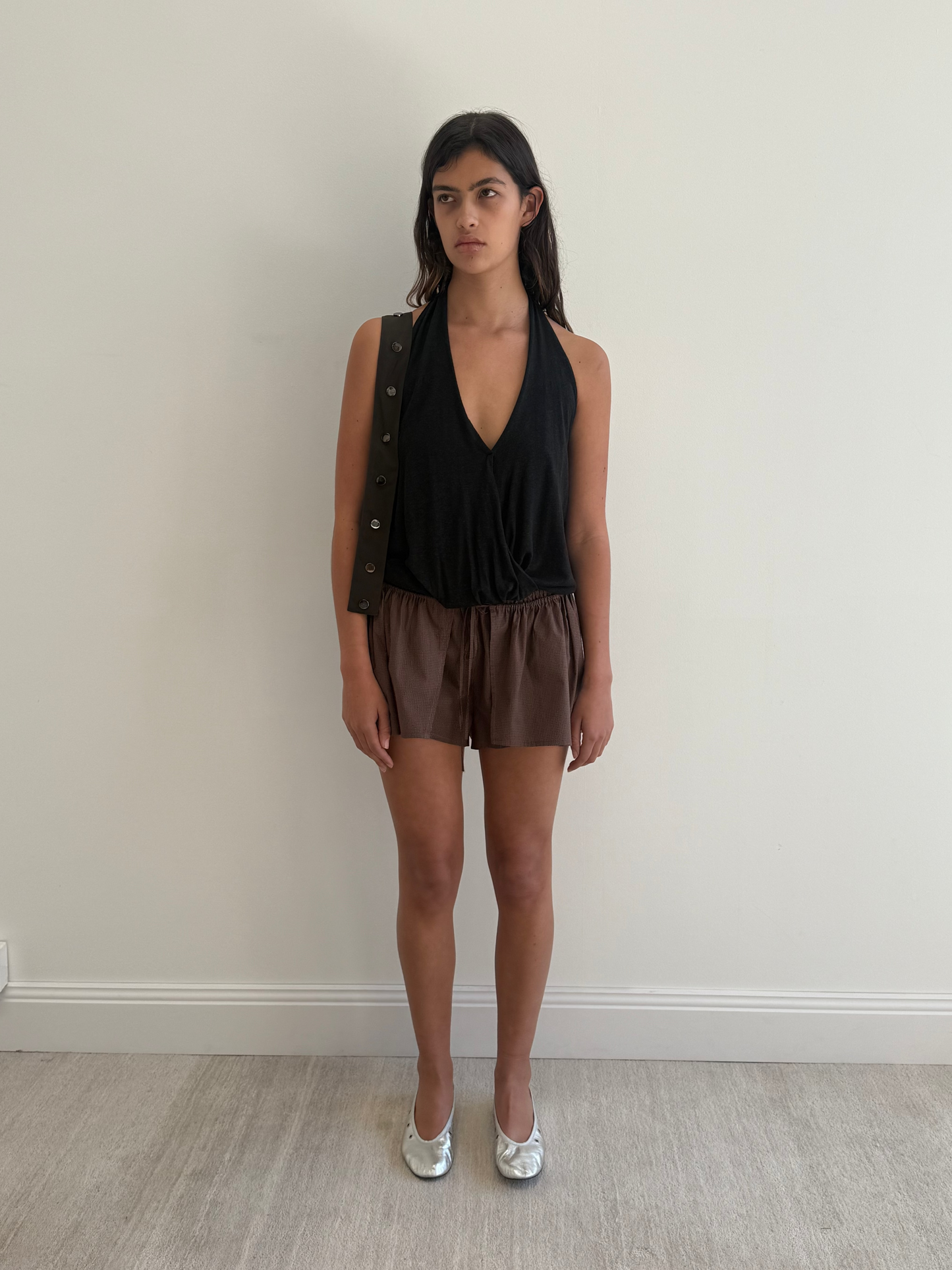 A woman with long dark hair stands against a plain white wall, wearing a sleeveless black top, the Skort Carob - Check by Deiji Studios, and silver flats. She looks neutral, arms resting at her sides.
