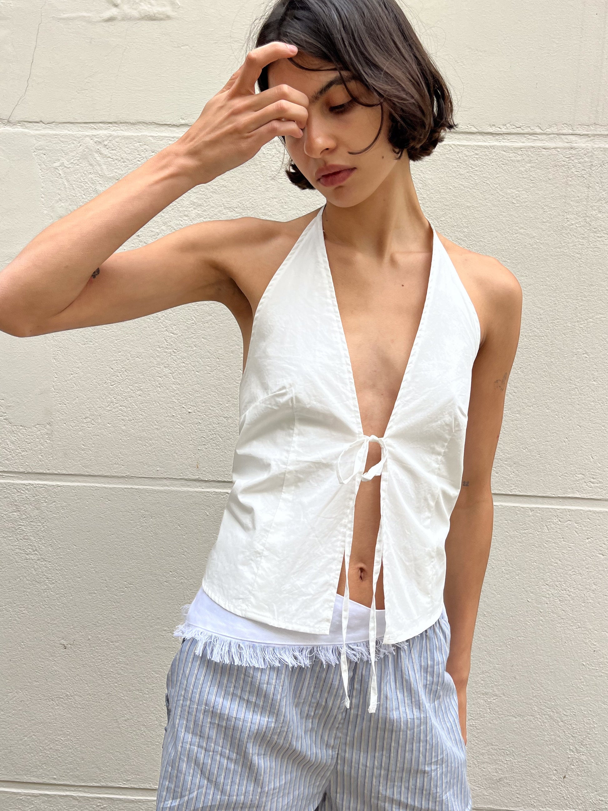 A woman with short dark hair wears the Open Halter Top - White, a sleeveless halter made from organic cotton, paired with light blue and white striped shorts, standing against a light wall and touching her forehead.