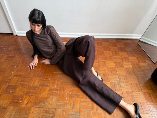 A woman with dark hair and a fringe reclines on a parquet floor propped on one arm, wearing the Linen Ease Trouser - Brown Black Stripe by Deiji Studios, dark brown fine-striped wide-leg linen trousers, with a matching brown striped knit top.