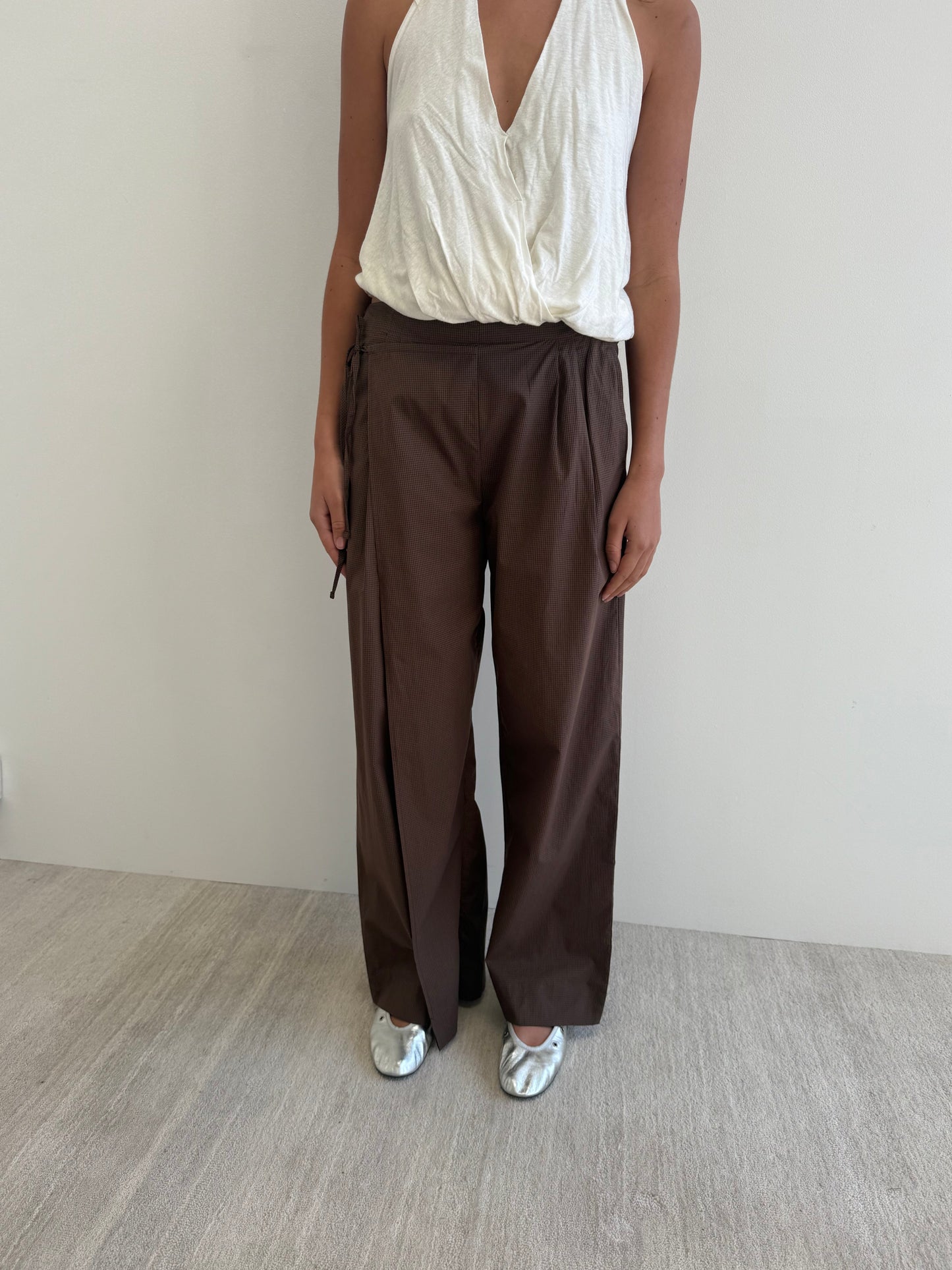 A person wearing a sleeveless white Deiji Studios wrap top, Double Layer Pant - Carob Check, and metallic silver shoes stands against a plain light-colored wall and floor. Their head is not visible in the image.
