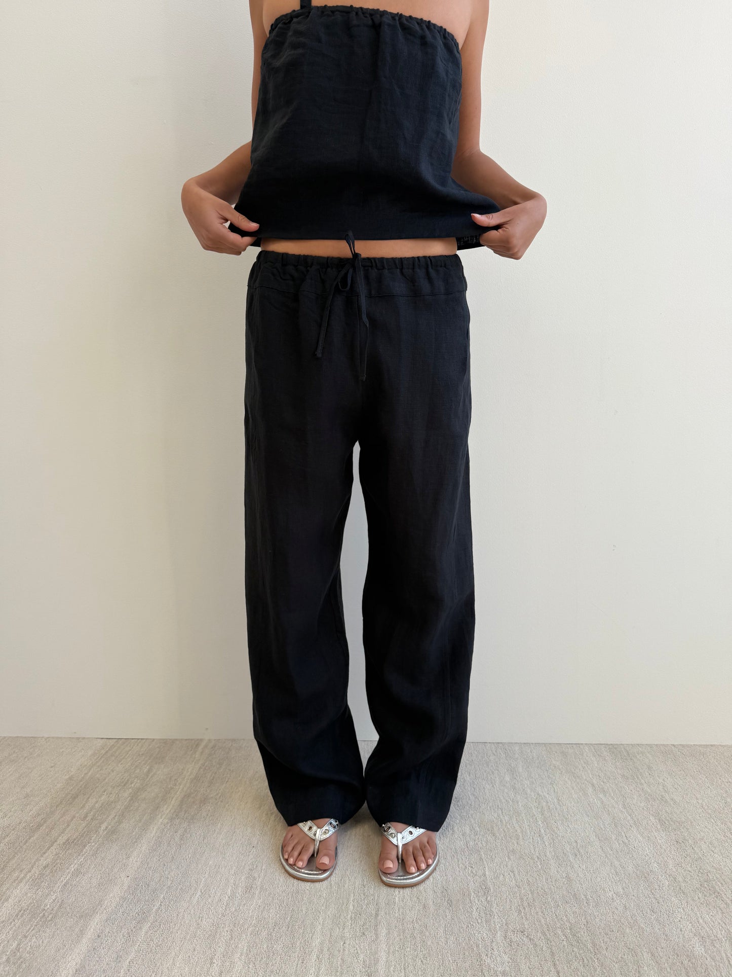 A person stands indoors against a plain wall wearing the Double Waist Linen Pant - Black and a matching sleeveless black top with silver sandals, slightly lifting the top to reveal the pants’ unique double waistband.