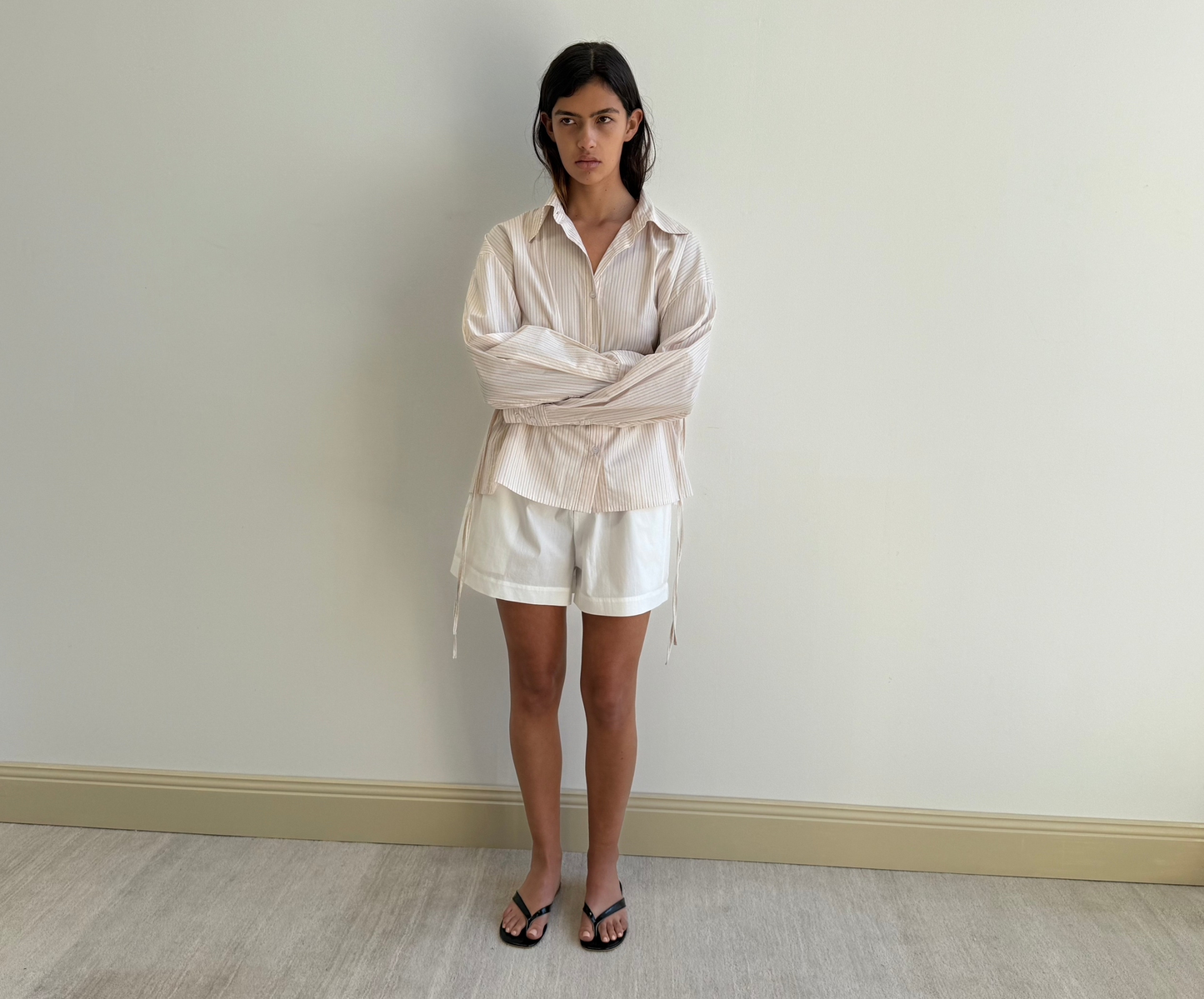 A woman with straight dark hair and a neutral expression stands indoors against a light wall, wearing the Deiji Studios Room Shirt - Dusty Stripe, loose white shorts, and black sandals. Her arms are relaxed by her sides.