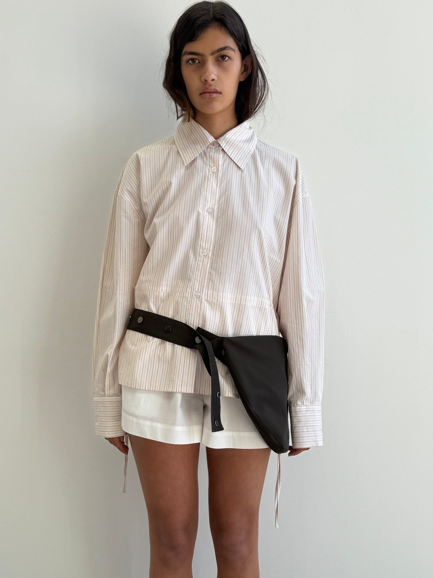 A woman with long dark hair and a neutral expression stands against a white background, wearing the Deiji Studios Room Shirt in Dusty Stripe, white shorts, and a black belt bag slung diagonally across her waist.