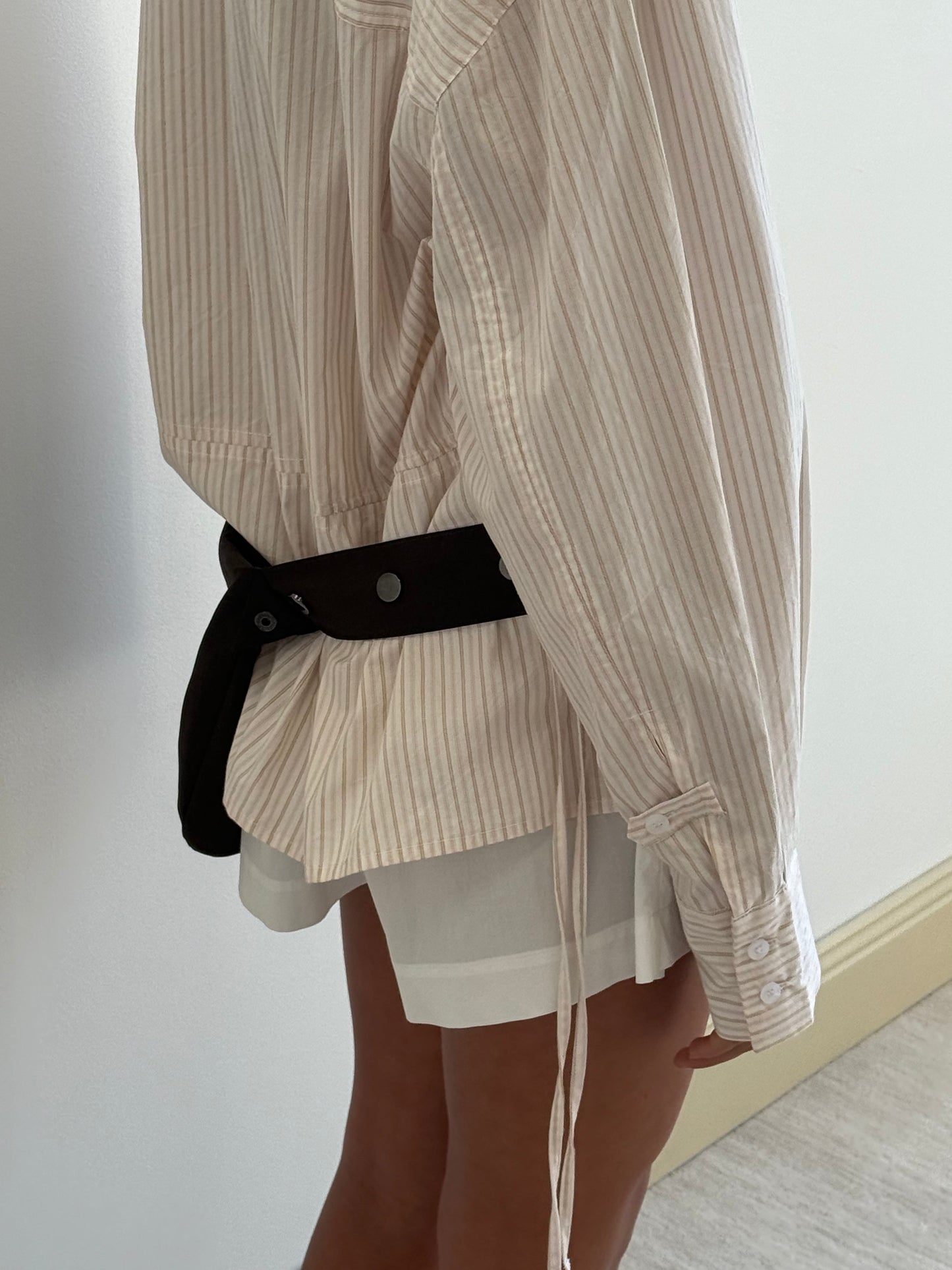 A person wearing the Room Shirt - Dusty Stripe stands sideways near a white wall, paired with white shorts and a loosely hanging black strap around their waist.