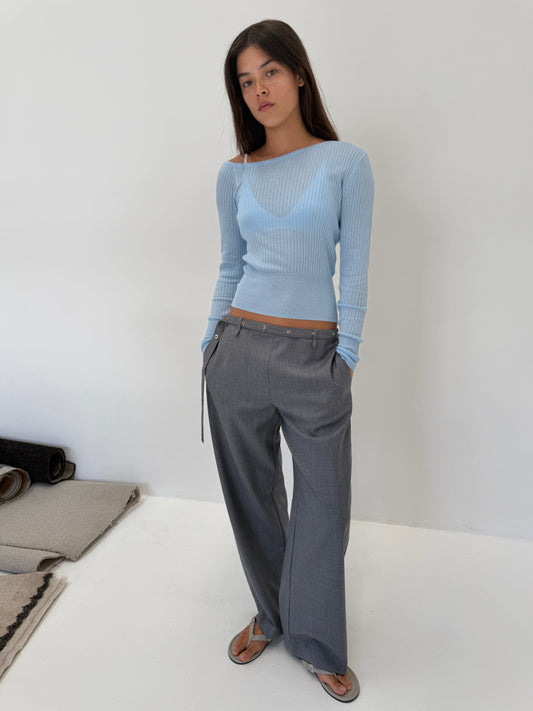 A woman with straight dark hair and a neutral expression stands indoors against a white wall, wearing the Shoulder Knit Top - Blue by Deiji Studios with gray wide-leg trousers and sandals. Her hands are in her pockets.