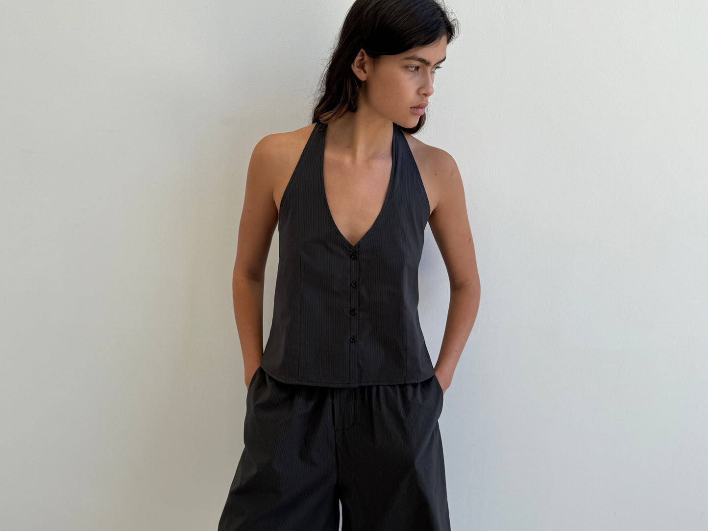A person wearing the Waist Halter - Black Stripe set from Deiji Studios, featuring a sleeveless, buttoned top and matching loose pants, stands against a white wall, looking to the side with hands in pockets.