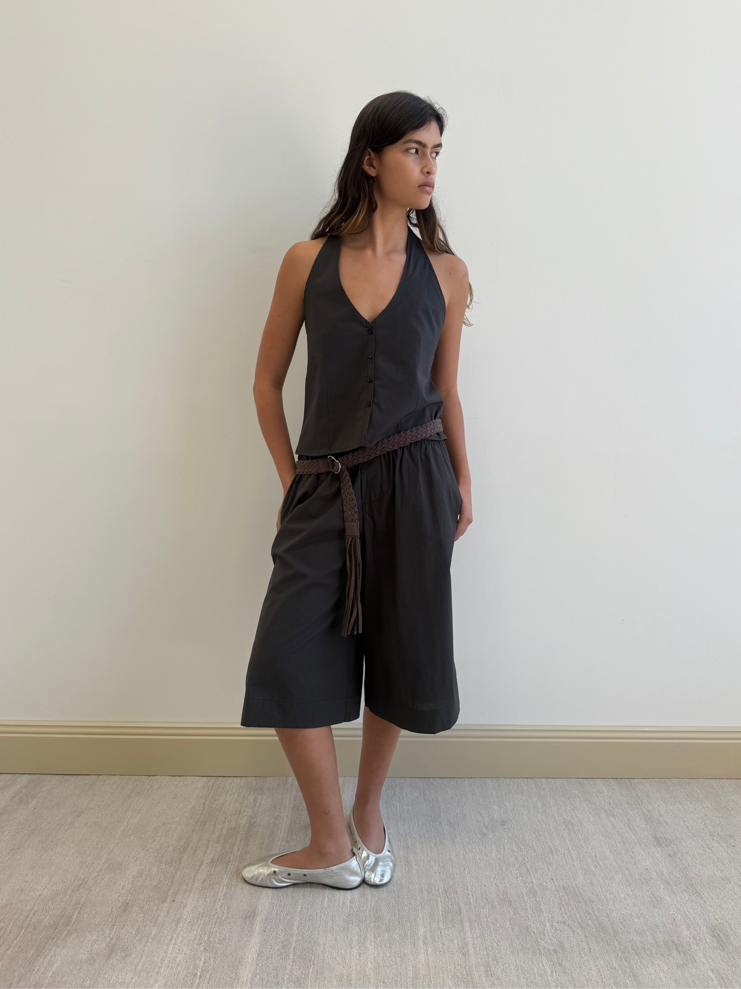 A woman stands indoors against a plain wall, wearing the Waist Halter - Black Stripe jumpsuit by Deiji Studios in organic cotton with a braided belt and silver flats. She looks to the side, hands in her pockets.