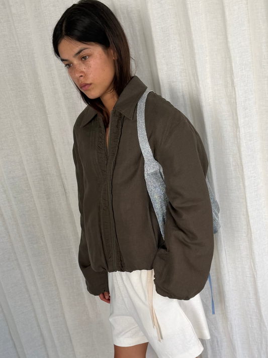 A woman with straight dark hair stands before white curtains, wearing the Pintuck Jacket - Khaki Stripe over a white skirt or shorts and a grey backpack, gazing downward with a neutral expression.