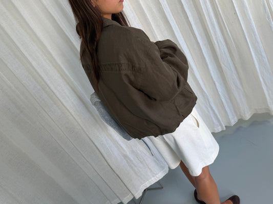 A woman stands indoors by white curtains, wearing a loose Deiji Studios Pintuck Jacket in Khaki Stripe over a white skirt, hair down and arms folded, with only her lower face visible.