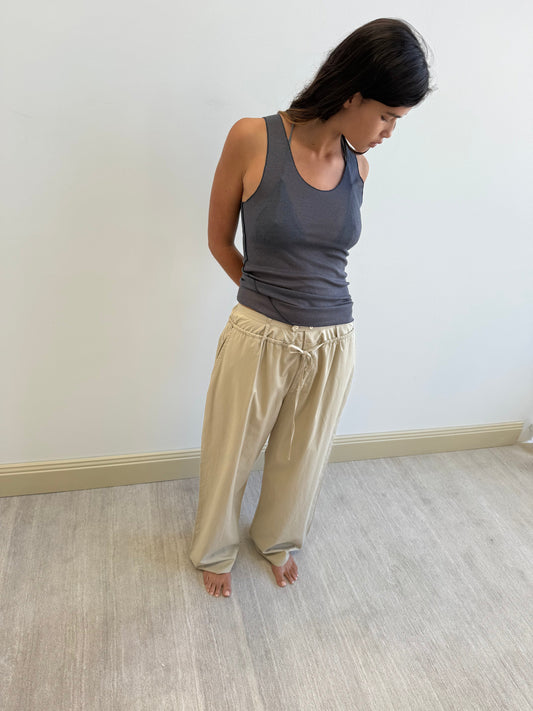 A woman with long dark hair stands barefoot on a light floor, wearing a gray tank and Deiji Studios’ Two Band Pant - Fawn, hands behind her back, looking down and slightly to the side.