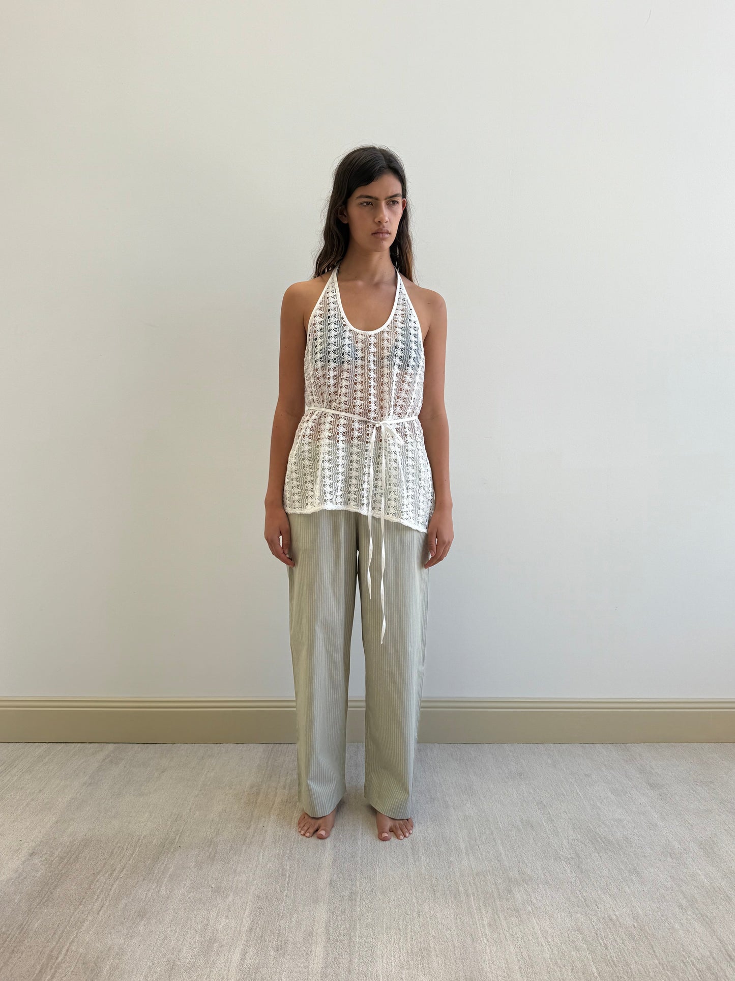 A woman with long dark hair and a neutral expression stands barefoot indoors against a plain wall, wearing loose light gray pants and Deiji Studios’ Thread Wrap Top in Off White, crafted from organic cotton.