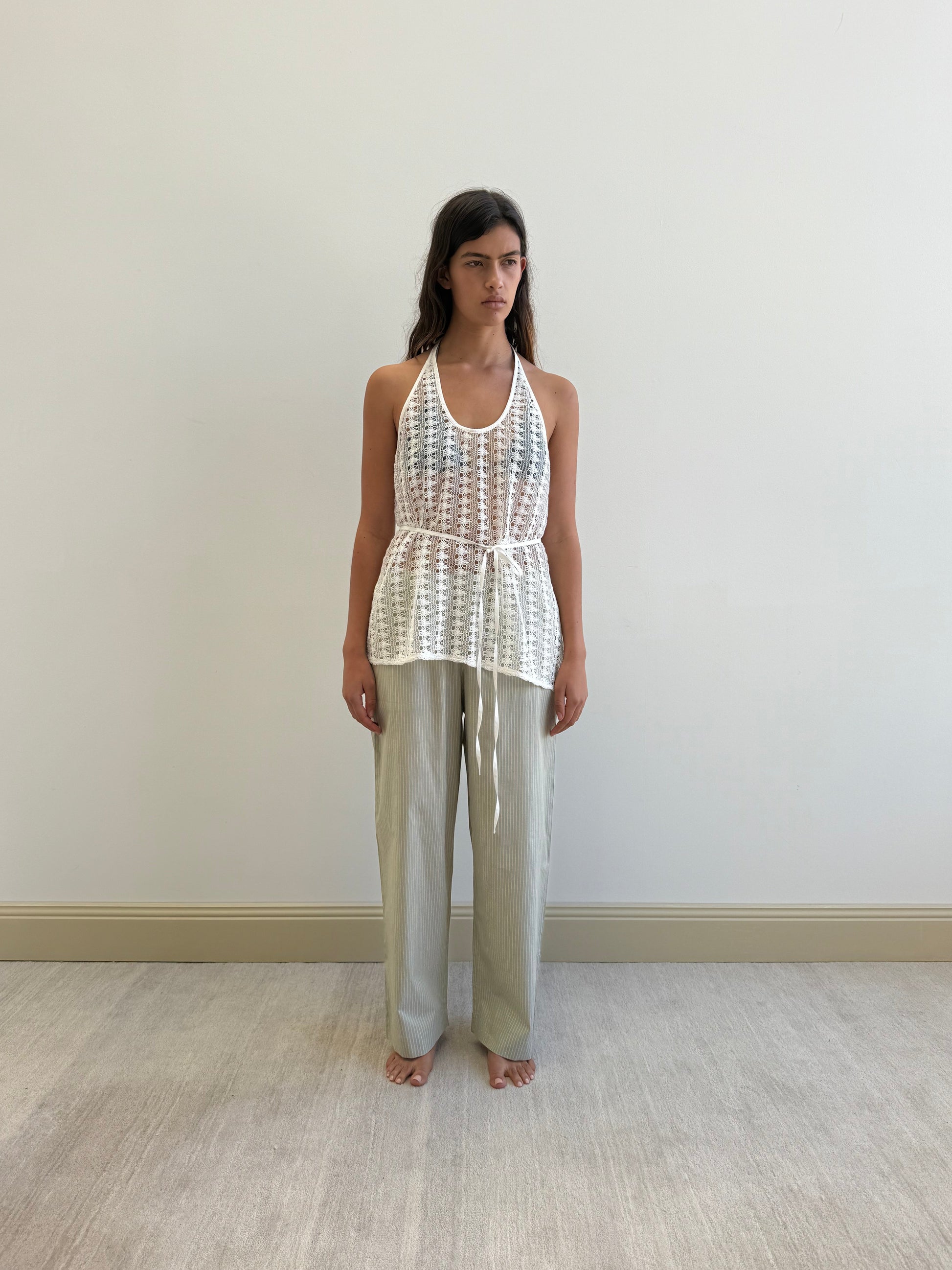 A woman with long dark hair and a neutral expression stands barefoot indoors against a plain wall, wearing loose light gray pants and Deiji Studios’ Thread Wrap Top in Off White, crafted from organic cotton.