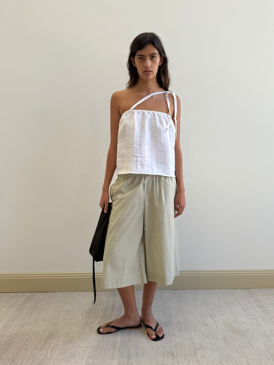 A woman stands indoors against a plain wall wearing the Long Lounger - Pistachio Stripe from Deiji Studios, styled with loose light-colored shorts, black flip-flops, and holding a black bag at her side.