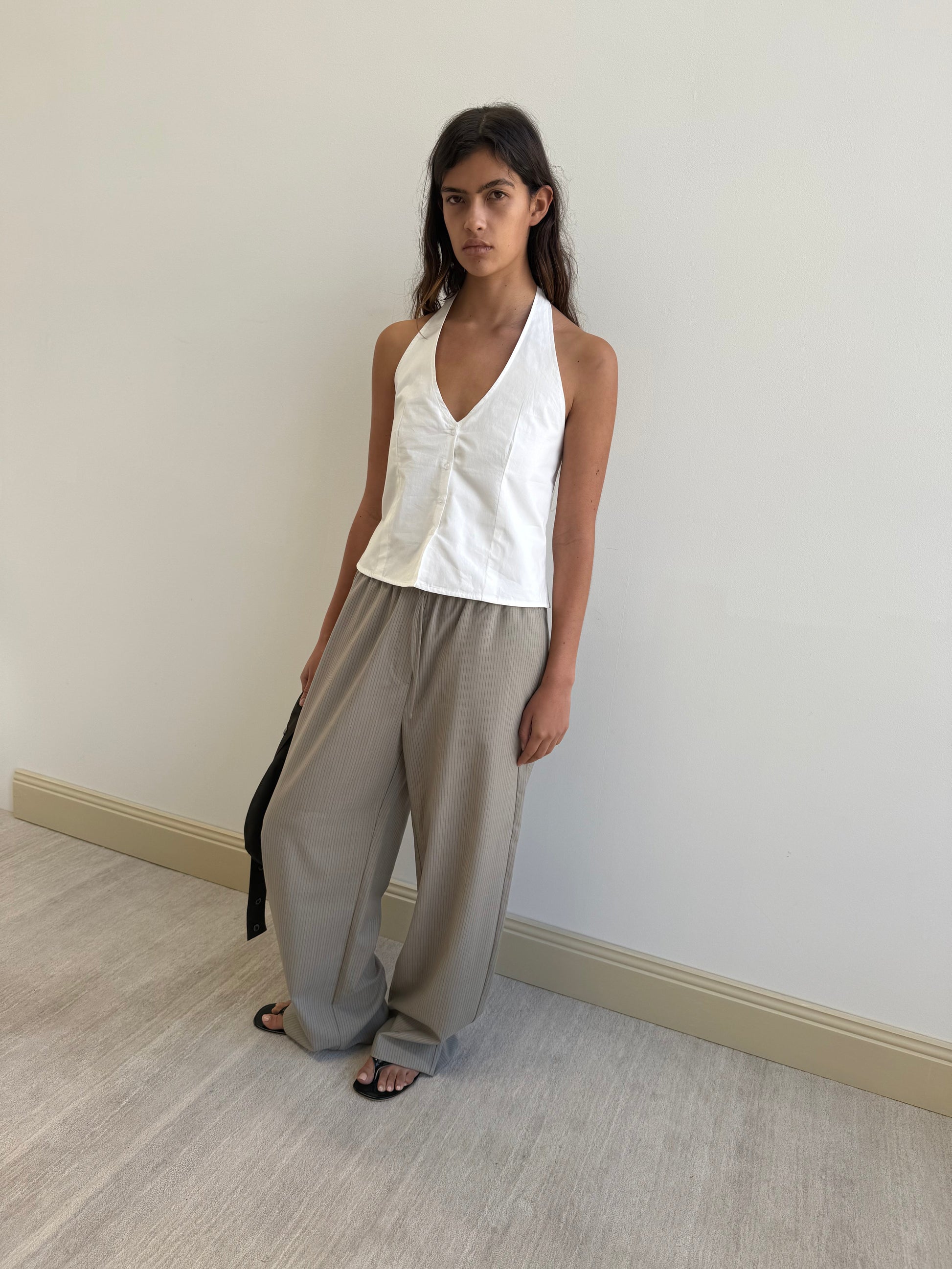 A woman stands indoors against a plain light wall, wearing the Waist Halter - White by Deiji Studios, loose gray trousers, and black sandals, holding a black strap in her left hand.