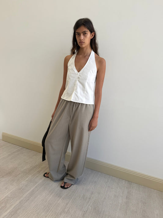 A woman stands indoors against a plain light wall, wearing the Waist Halter - White by Deiji Studios, loose gray trousers, and black sandals, holding a black strap in her left hand.