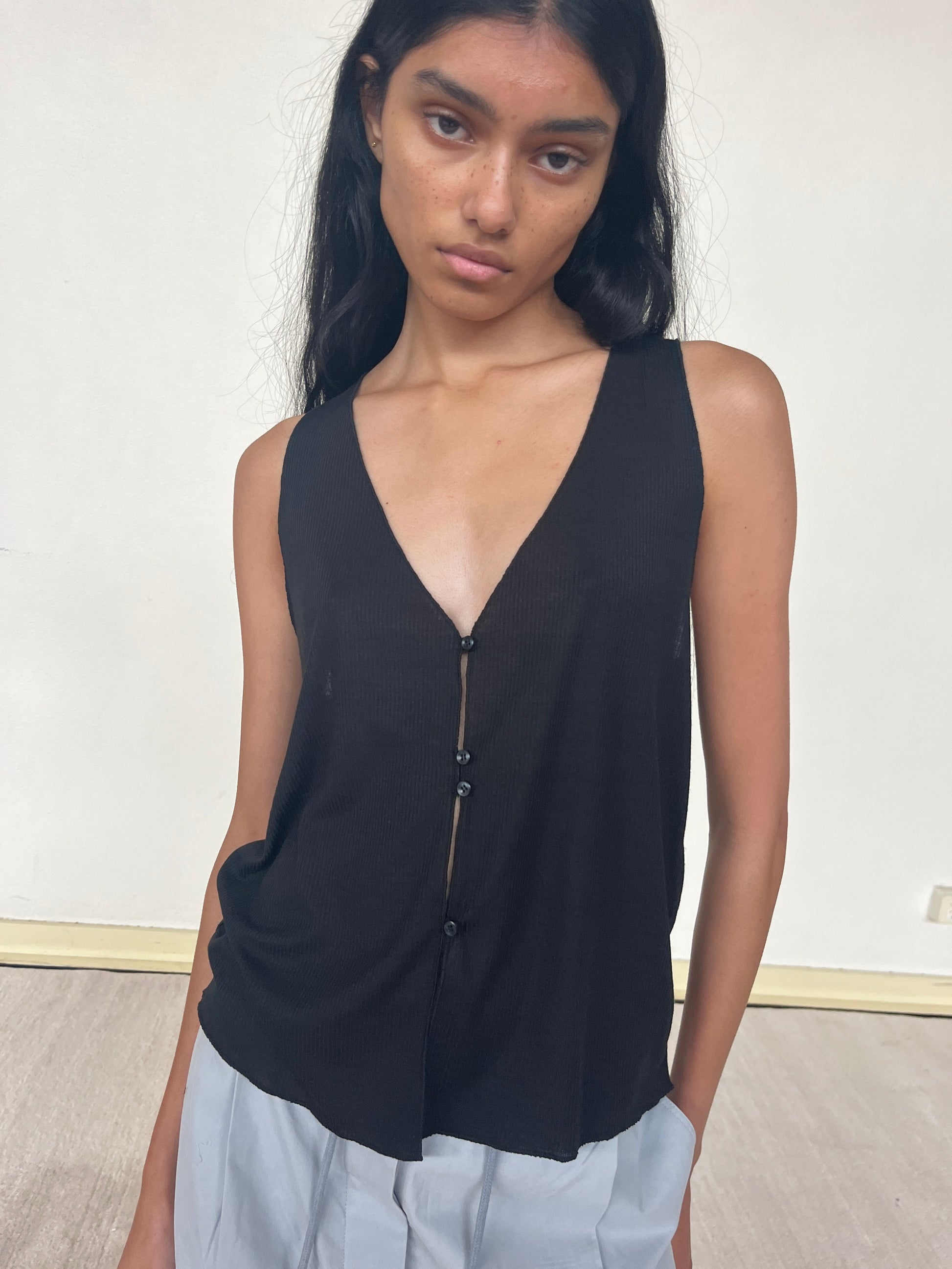 A woman with long dark hair wears the Button Up Knit Tank, a sleeveless black top with a deep V-neck and buttons, paired with light gray pants. She stands against an off-white wall, posing naturally with a neutral expression.