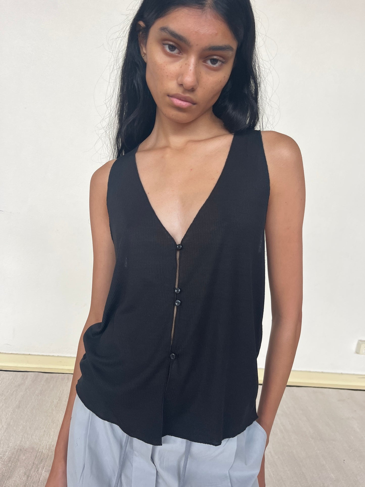 A woman with long dark hair wears the Button Up Knit Tank—a sleeveless black V-neck top with buttons—paired with light gray pants, standing indoors against a plain white wall with a neutral expression and relaxed posture.