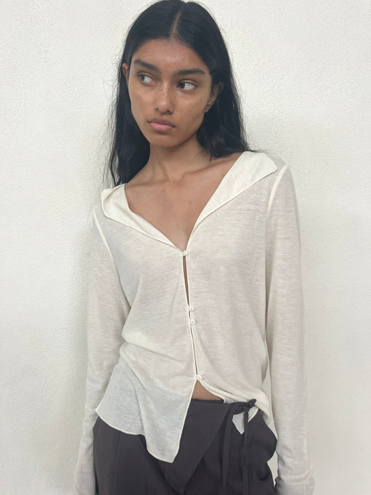 A woman with long dark hair stands against a white wall, wearing the cream-colored Open Long Sleeve Top and brown pants. She looks slightly to the side with a neutral expression.