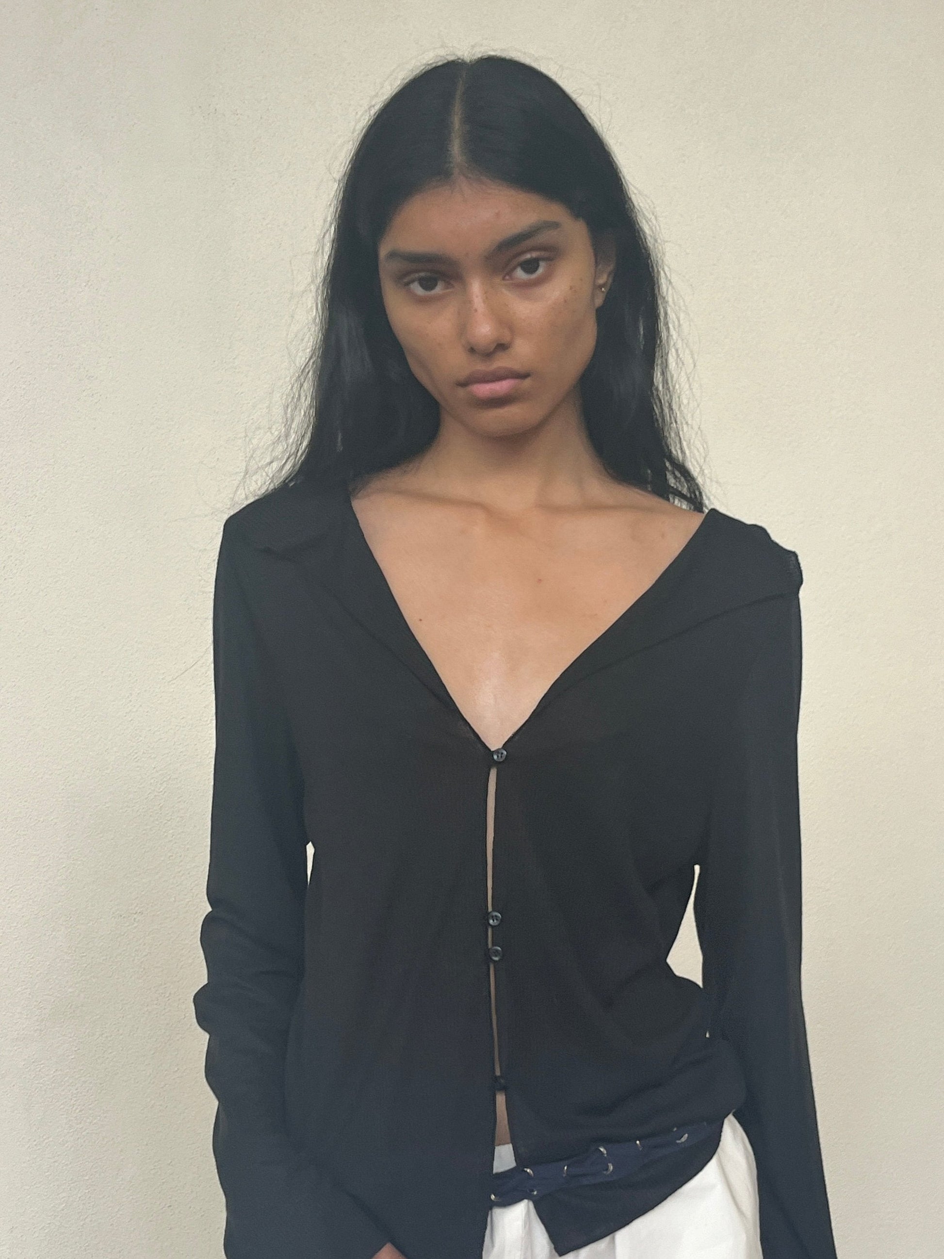 A person with long black hair models the Open Long Sleeve Top—a black, deep V-neck buttoned shirt—paired with white pants against a plain light background, gazing at the camera with a neutral expression.