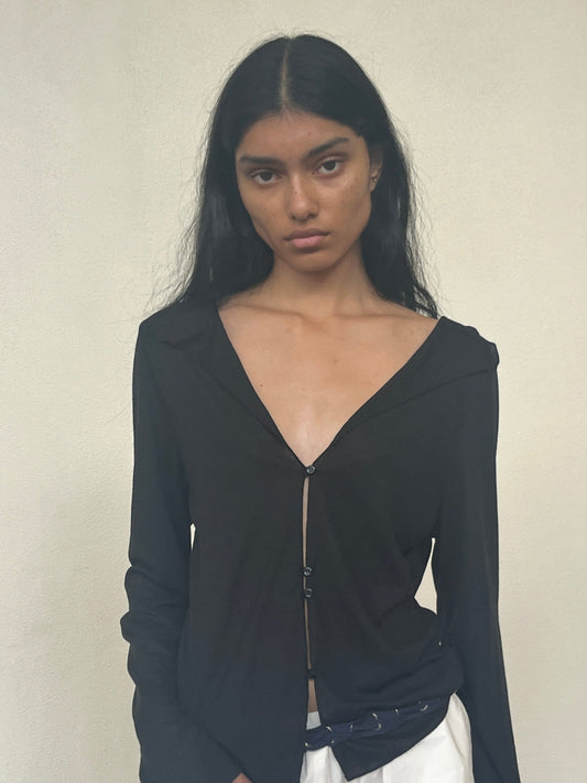 A person with long black hair models the Open Long Sleeve Top—a black, deep V-neck buttoned shirt—paired with white pants against a plain light background, gazing at the camera with a neutral expression.