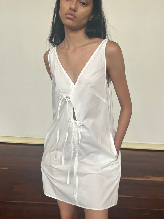 A person stands indoors, hands in pockets, wearing the Open Tie Mini Dress—a sleeveless, loose-fitting white dress with a deep V-neck and two front ties—posed against a plain background with wooden flooring.