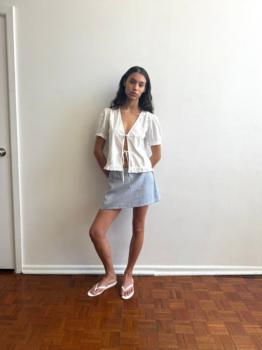A woman stands indoors by a plain white wall, wearing Deiji Studios’ Babylock Skirt in Blue Stripe with a white tie-front blouse and sandals. Her long hair is down as she gazes neutrally at the camera.