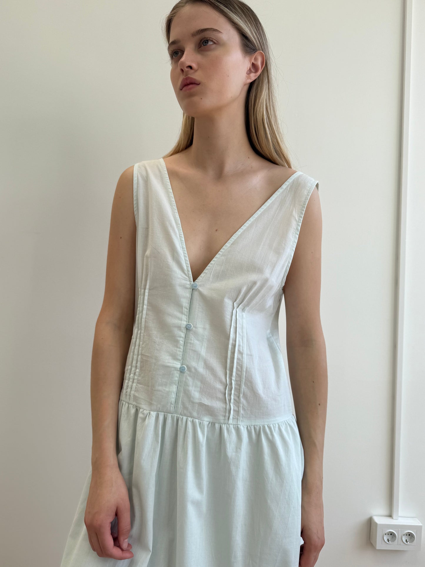 A woman with long blond hair stands against a plain light wall, wearing the Pintuck Dress - Mint from Deiji Studios, a sleeveless organic cotton dress with a deep V-neck and front buttons. She gazes to the side with a neutral expression.