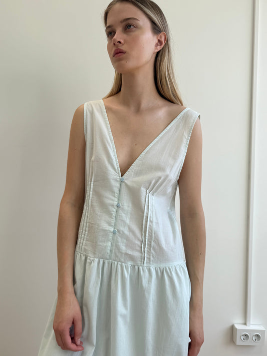 A woman with long blond hair stands against a plain light wall, wearing the Pintuck Dress - Mint from Deiji Studios, a sleeveless organic cotton dress with a deep V-neck and front buttons. She gazes to the side with a neutral expression.