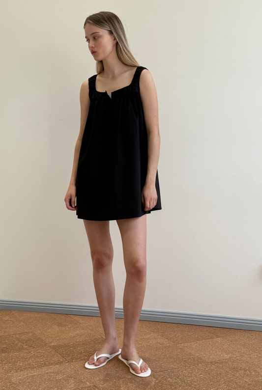 A woman with long blonde hair stands indoors against an off-white wall and cork floor, wearing Deiji Studios’ Yoke Mini Dress - Black and white flip-flops, glancing to the side.