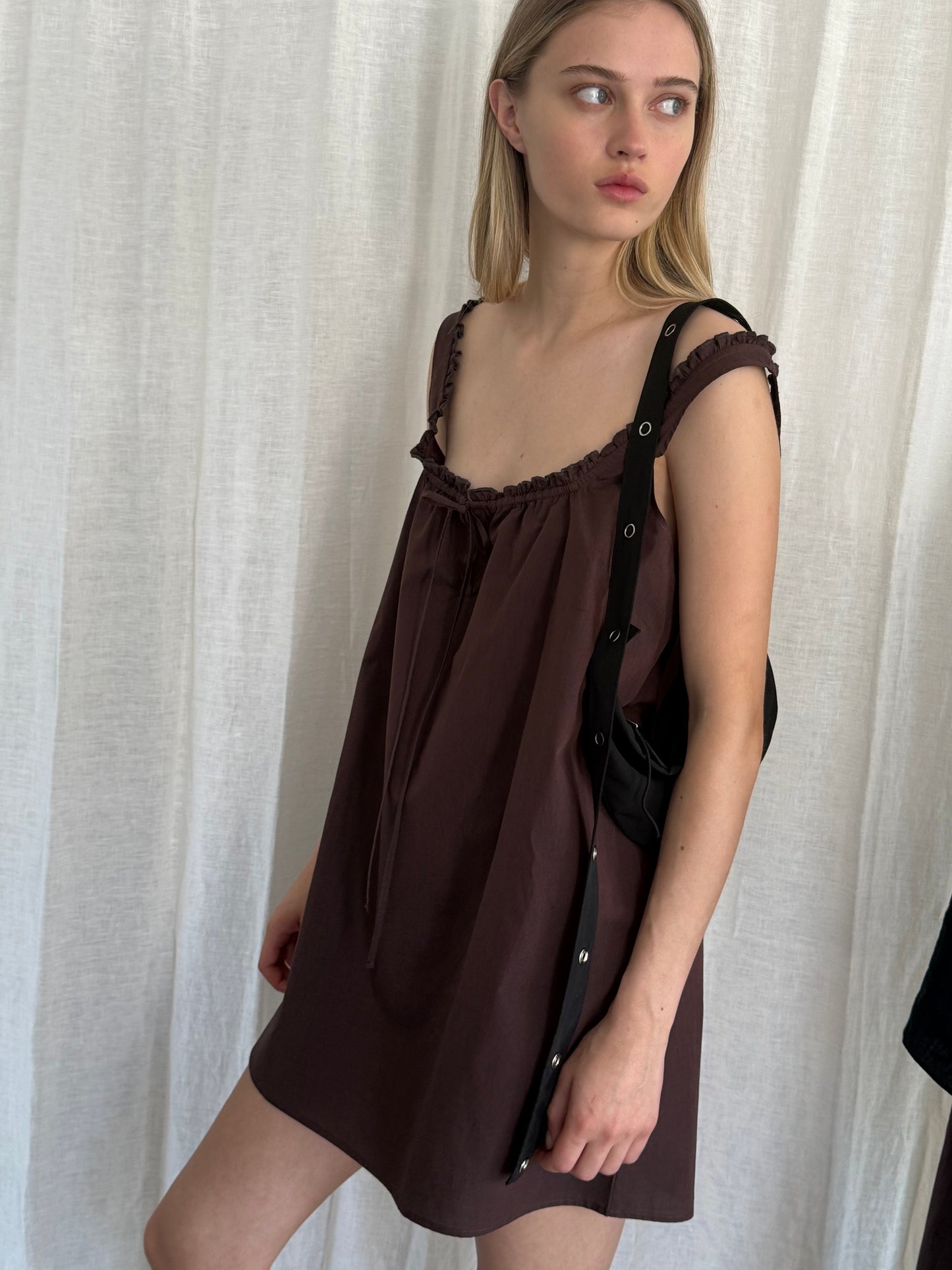 A young woman with long blonde hair stands in front of a white curtain, looking right. She wears a loose, sleeveless Deiji Studios Button Frill Mini Dress in Umber and carries a black shoulder bag.