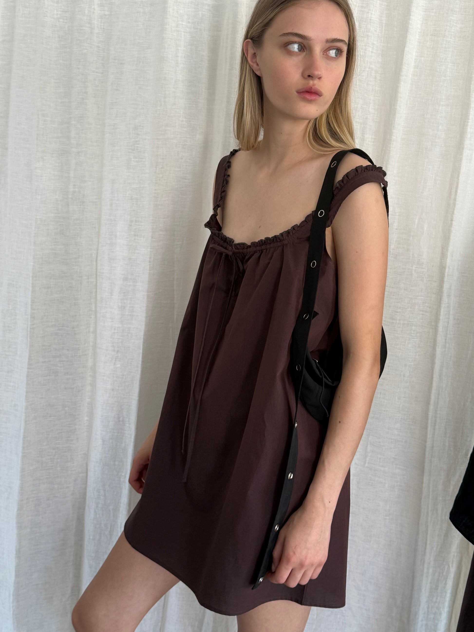 A young woman with long blonde hair stands in front of a white curtain, looking right. She wears a loose, sleeveless Deiji Studios Button Frill Mini Dress in Umber and carries a black shoulder bag.