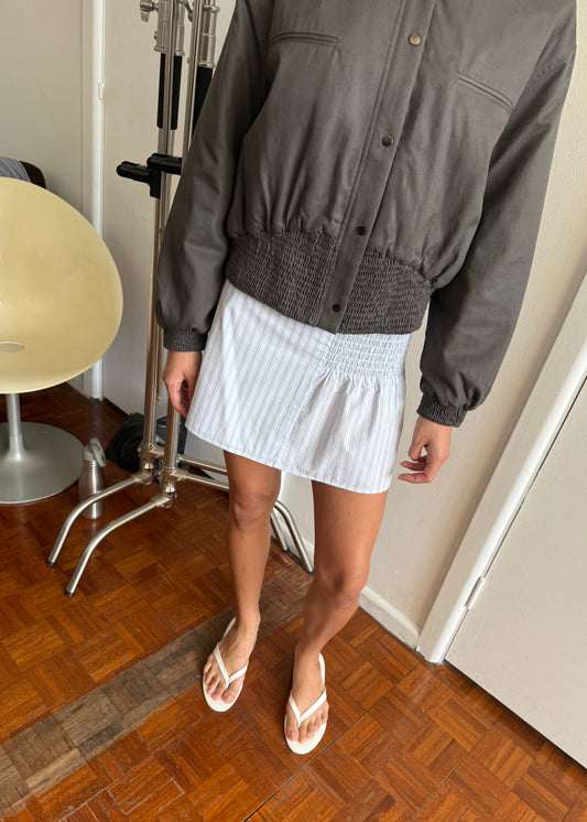 A person stands indoors on a wooden floor, wearing a gray jacket and the Deiji Studios Shirring Skirt in Mineral Stripe with white flip-flops. A cream chair and some metal equipment can be seen in the background.