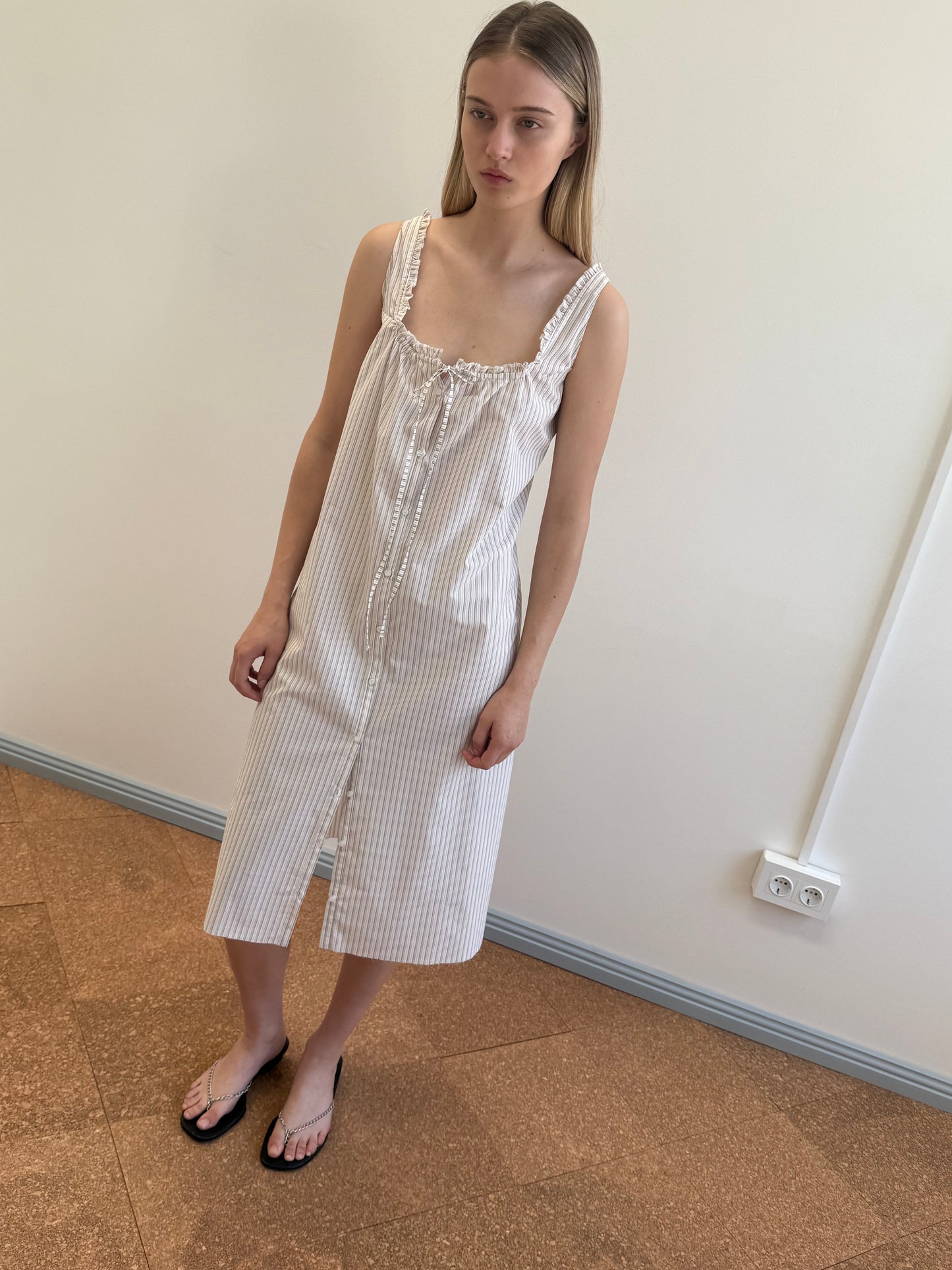 A model wears the Button Sweep Dress—a white and grey striped midi sundress with ruffled straps, drawstring neckline, and front slit—paired with black chain-detail flip-flops.