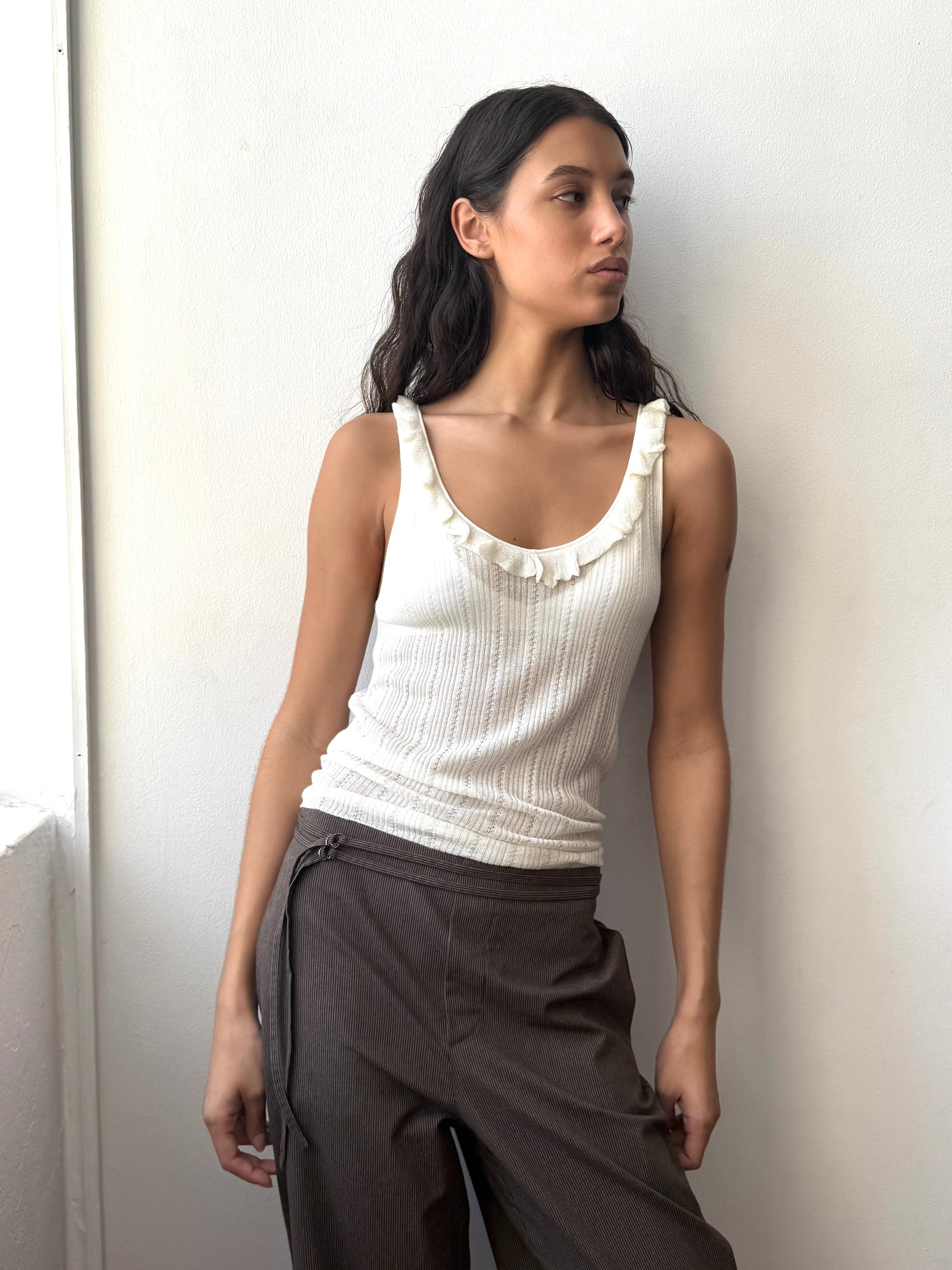 A woman with long dark hair wears the Neck Ruffle Knit - White top and brown pants, standing against a plain white wall and looking to her left.