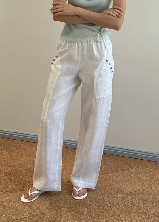 The Angle Pocket Pant - White by Deiji Studios features a wide-leg linen design, elastic waistband, side buttons, and patch pockets. Styled here with a mint green knit top and white flip-flops.