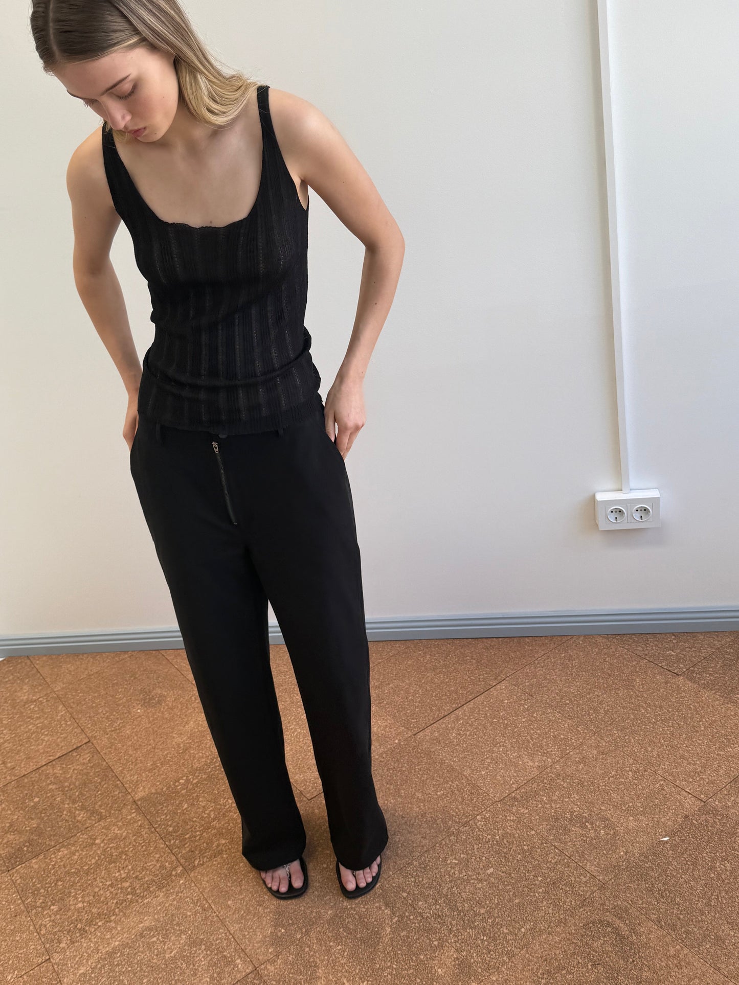 A woman with straight, light brown hair looks down while wearing the Deiji Studios Open Work Tank in black, paired with black pants and sandals. She stands indoors against a plain wall with an electrical outlet in view.