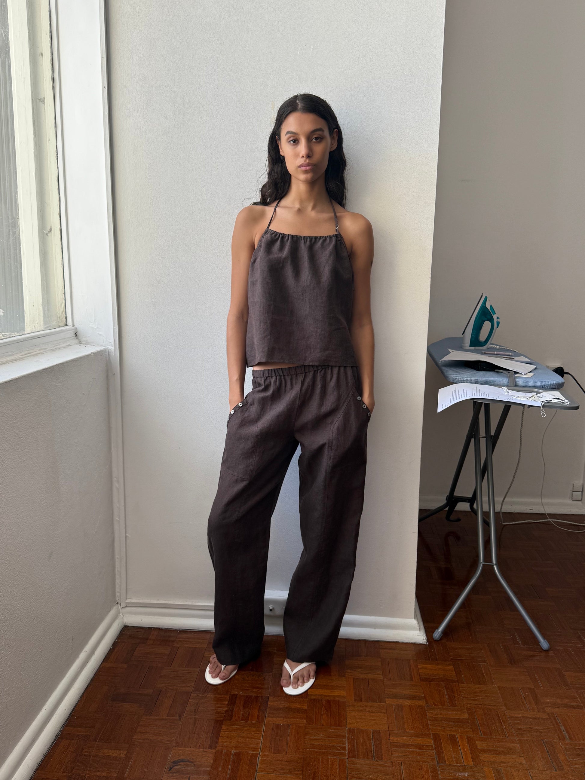 A woman stands indoors by a window, wearing the Angle Pocket Pant in dark brown with a matching sleeveless top and white sandals. She has her hands in her pockets. An iron and ironing board are visible in the background.