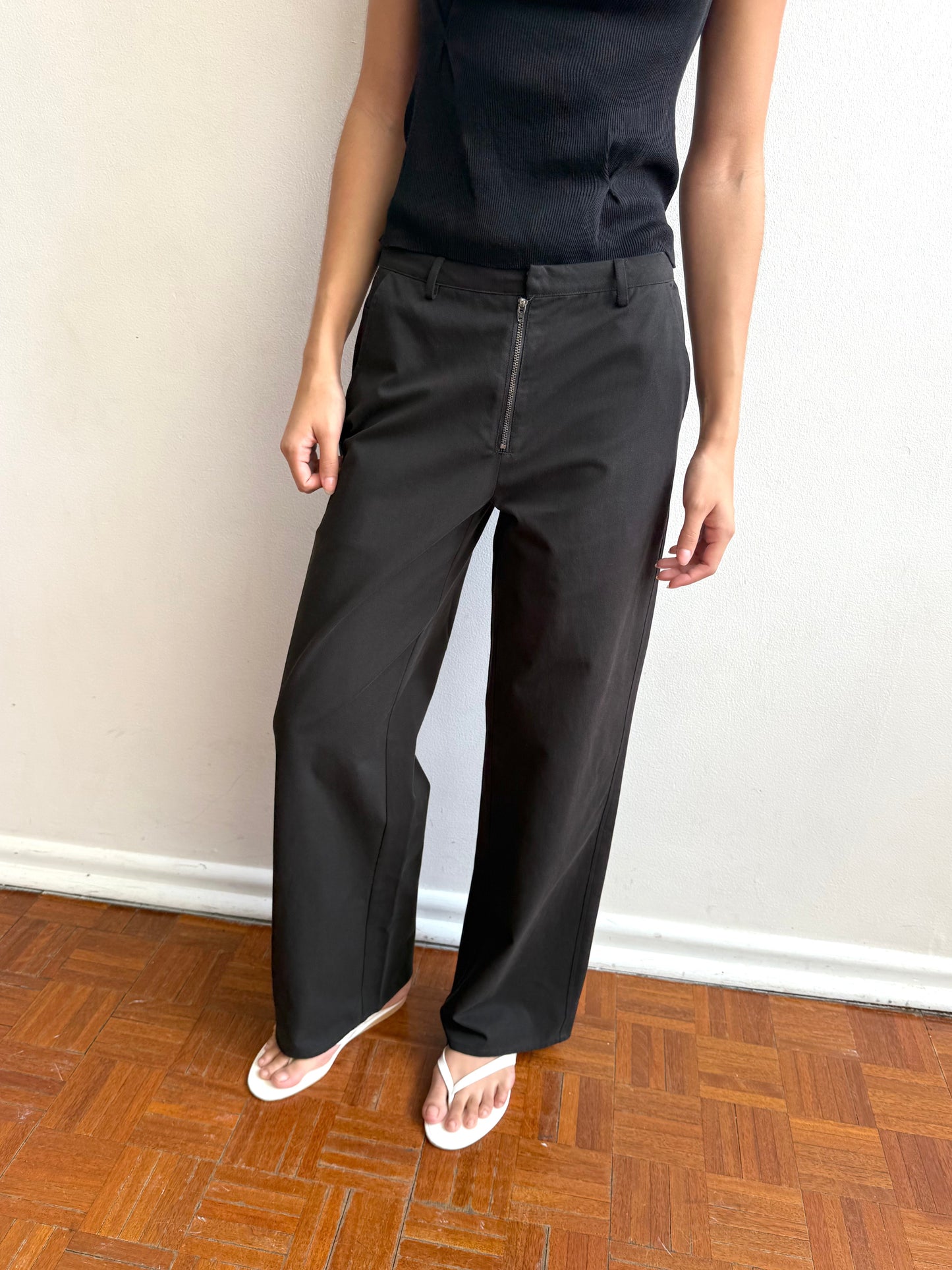A person in a sleeveless black top, Zip Pant - Black (organic cotton, straight leg), and white sandals stands by a light wall and wooden floor. The face is not visible.