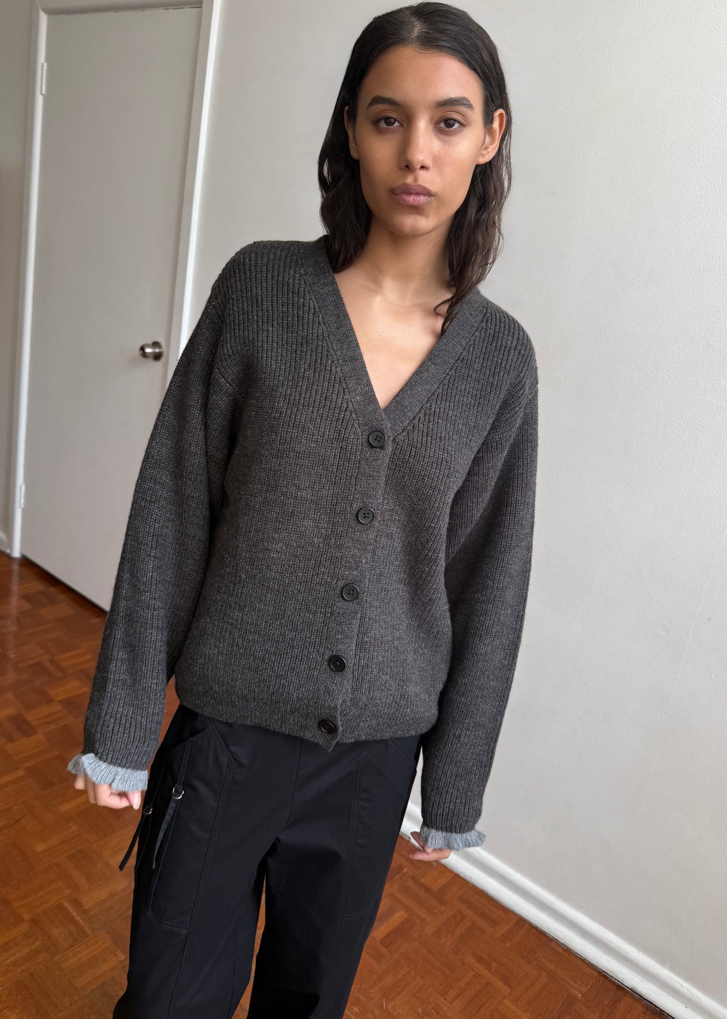 A woman with dark hair wears the Flounce Cardigan - Charcoal and black pants, standing indoors on a wooden floor against a white wall. She gazes calmly at the camera, embodying minimalist Deiji Studios style.