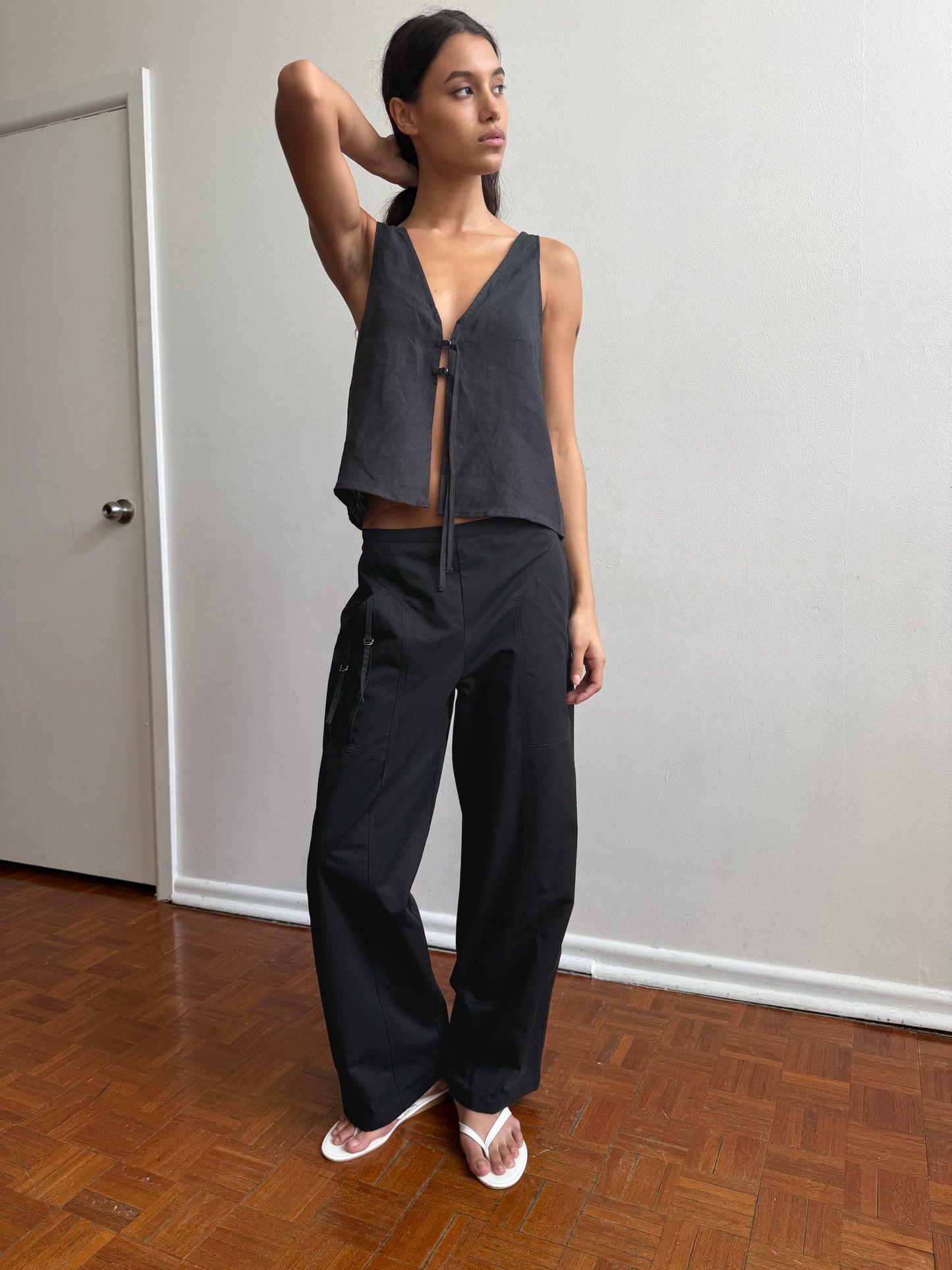 A woman poses indoors on a wooden floor, wearing a sleeveless black top, the Adjust Pocket Pant - Black by Deiji Studios in organic cotton, and white sandals. She stands in front of a plain white wall with one hand behind her head, gazing to the side.
