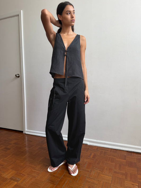 A woman poses indoors on a wooden floor, wearing a sleeveless black top, the Adjust Pocket Pant - Black by Deiji Studios in organic cotton, and white sandals. She stands in front of a plain white wall with one hand behind her head, gazing to the side.