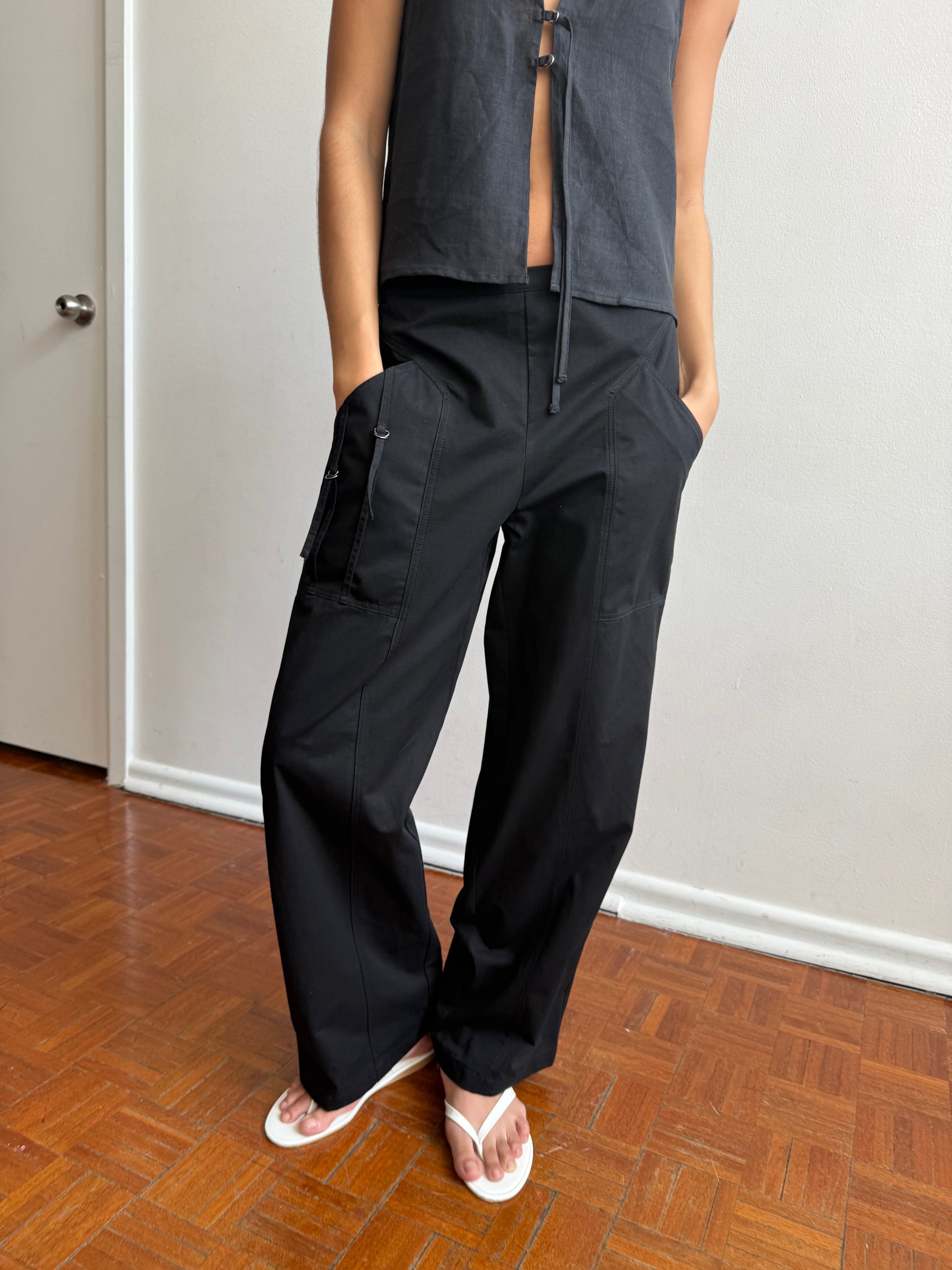 Indoors on a wooden floor, someone wearing Deiji Studios' Adjust Pocket Pant - Black and a sleeveless black top adjusts the pants' pocket details. They pair the look with white flip-flops; their head is not shown in the image.