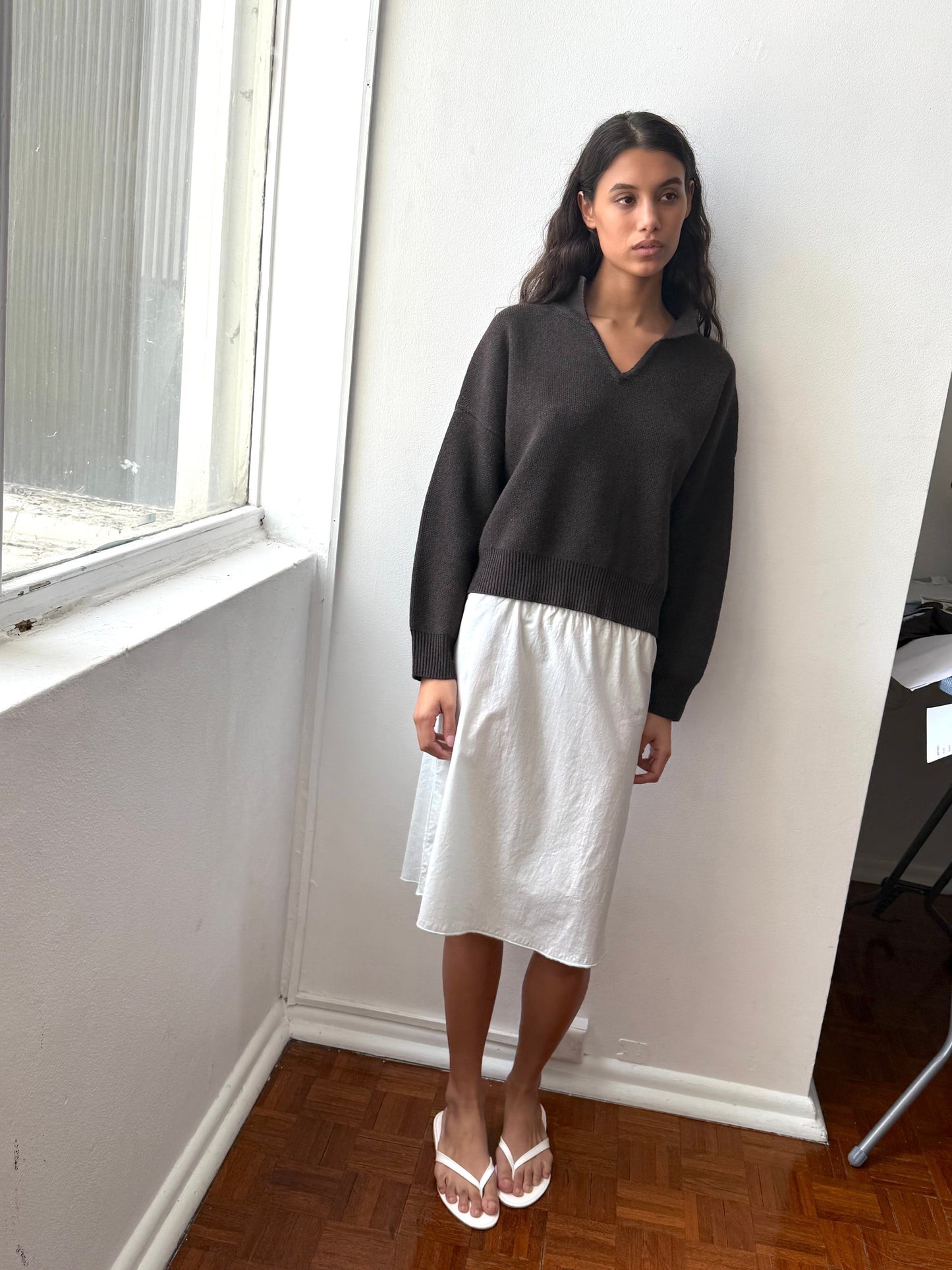 A woman stands indoors by a window, wearing a dark long-sleeve sweater, the Deiji Studios Simple Skirt - Mint in organic cotton, and white flip-flops. She has long, wavy dark hair and gazes to the side with a neutral expression.