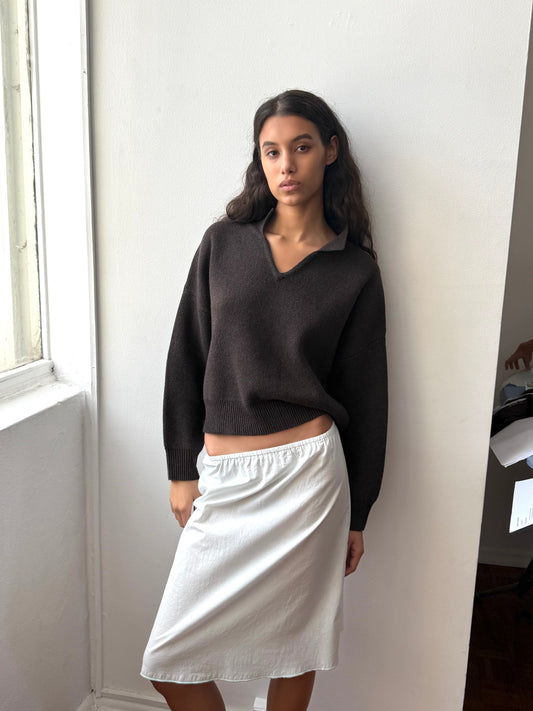 A woman stands indoors by a white wall and window, wearing a dark long-sleeve sweater and the Simple Skirt - Mint from Deiji Studios, looking calmly at the camera with one hand by her side.