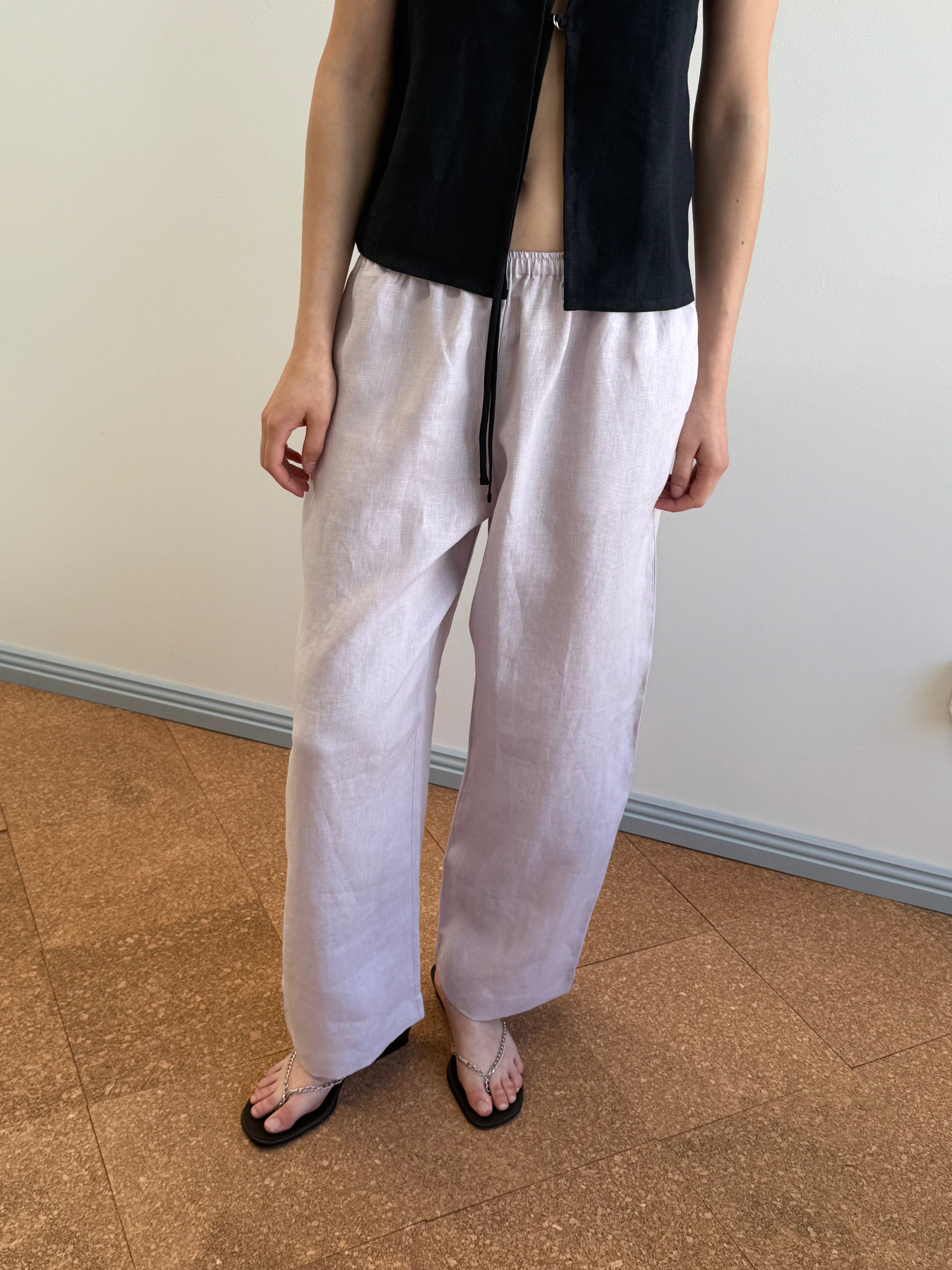 A person indoors wears the Linen Ease Trouser—loose-fitting white linen pants—paired with a black sleeveless top and black flip-flops. Only the lower half of their face and body are visible against a plain, light-colored background.