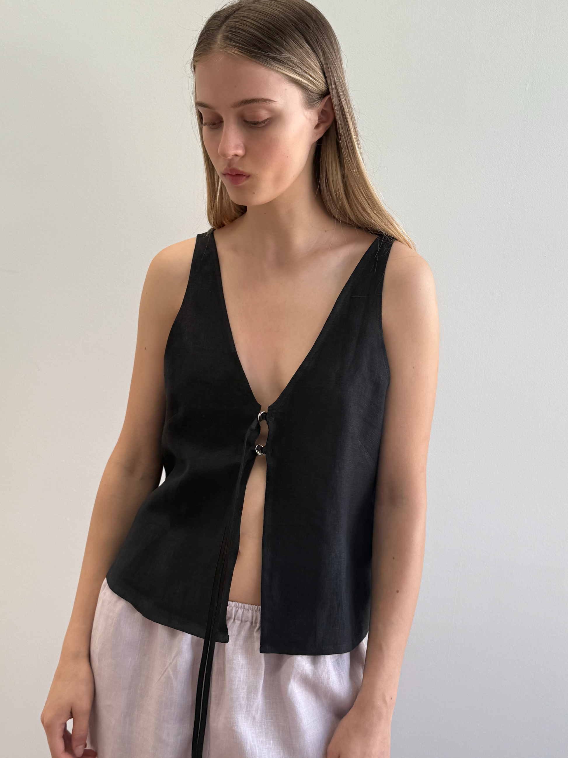 Model wears the Two Part Top—a black linen sleeveless top with a deep V-neckline and single ring-clasp front closure—paired with white linen wide-leg trousers, set against a light grey background.